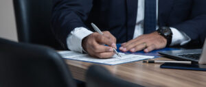 Homem com roupa social sentado em uma mesa assinando papéis